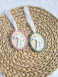 May include: Two Easter-themed wooden ornaments. One is framed in pink, the other in green, both with a bunny silhouette and a name. Each ornament has a white ribbon for hanging. The names "Rosie" and "Noah" are written in cursive.